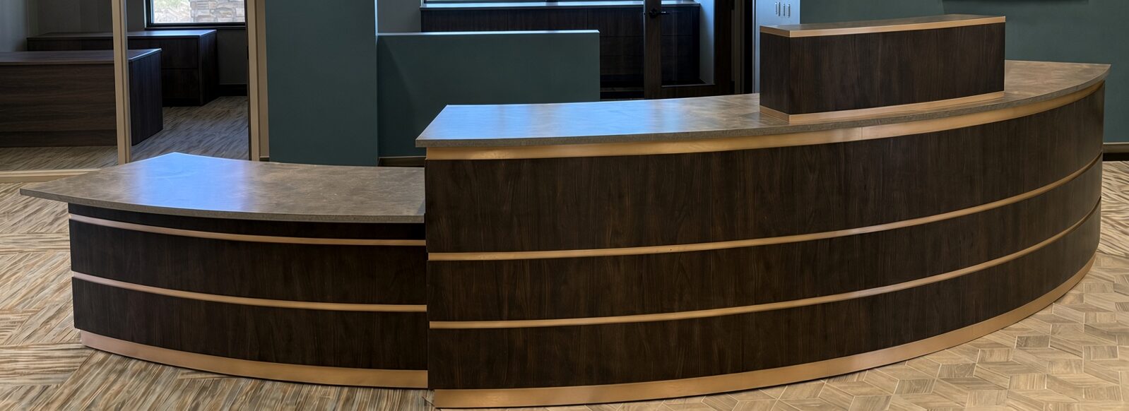 Curved reception desk with dark walnut-finish front, bronze-tone accent bands and gray stone-look top