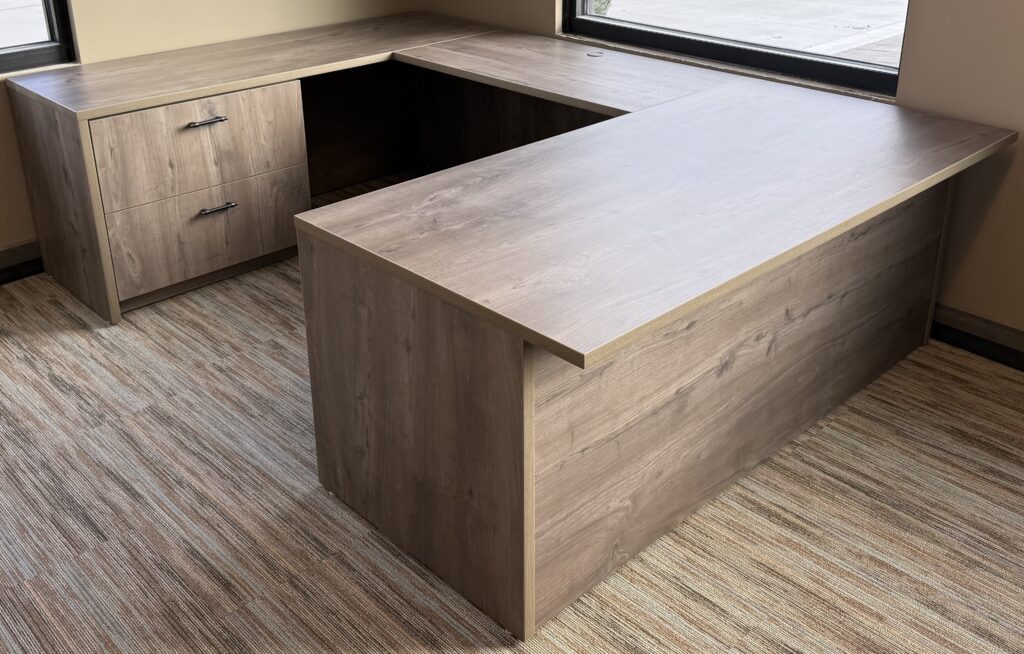 U-shaped desk with two-drawer lateral file in gray-brown oak-look finish beside office windows