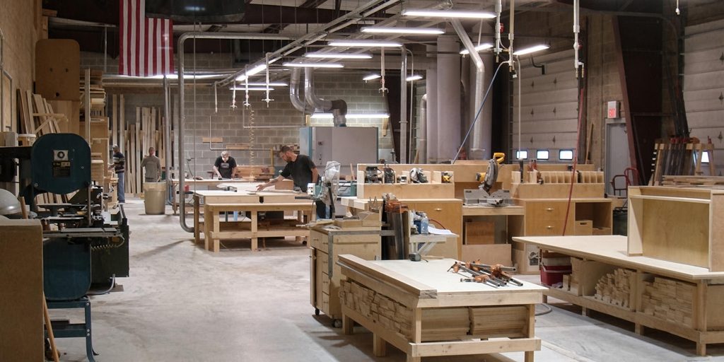 Cabinet shop floor with light-wood workbenches, casework in progress, power tools and overhead dust collection ducting